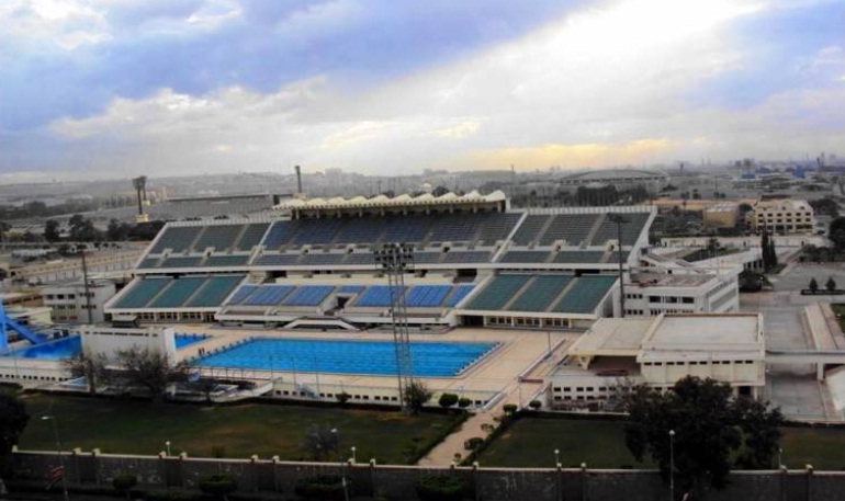 Luftaufnahme eines Schwimmstadions mit Tribünen und einem Außen-Wettkampfbecken.