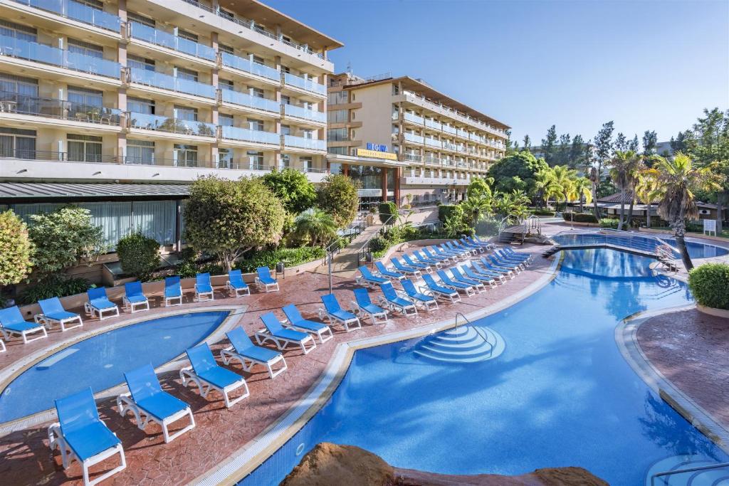 Außenpoolbereich mit großem, geschwungenem Becken, Sonnenliegen, Palmen und den Hotelgebäuden im Hintergrund.