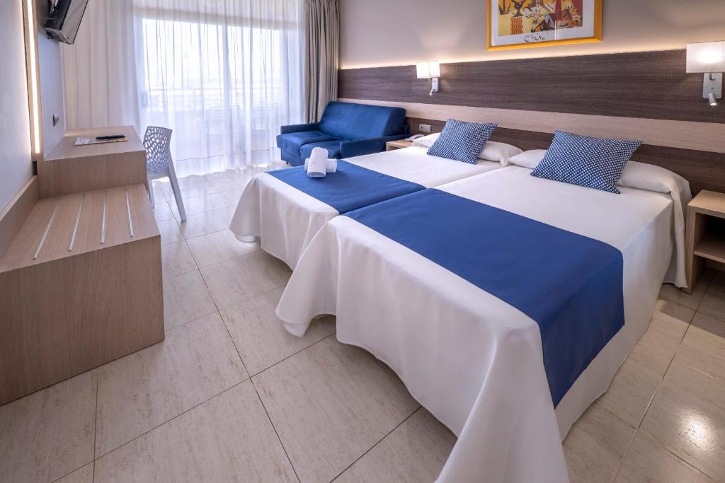 Geräumiges Zimmer mit zwei Betten, blauem Bettläufer, Sofa-Sitzbereich, Schreibtisch mit TV und Balkontüren mit Vorhängen.