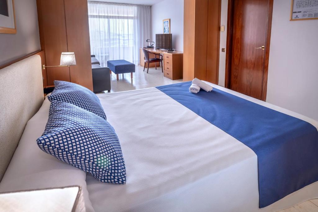 Schlafzimmeransicht mit Doppelbett, Schreibtisch und TV sowie Sitzbereich nahe dem Balkonfenster.