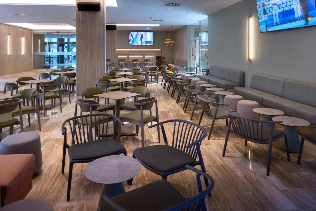 Moderner Hotel-Lounge-/Barbereich mit vielen Tischen und Stühlen, Sofas entlang der Wand, TVs und kleiner Bartheke.