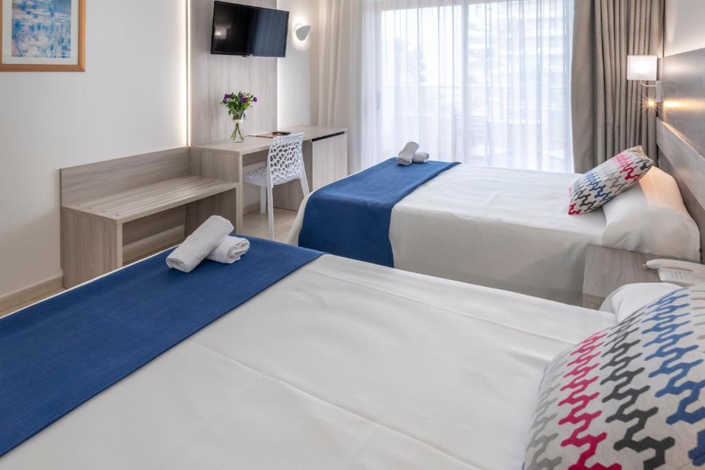 Zweibettzimmer mit zwei Einzelbetten, blauen Läufern, Wand-TV und Schreibtisch am Fenster.