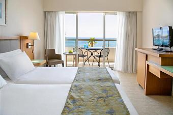 Guest room with sea-view balcony, twin beds, TV and desk area.