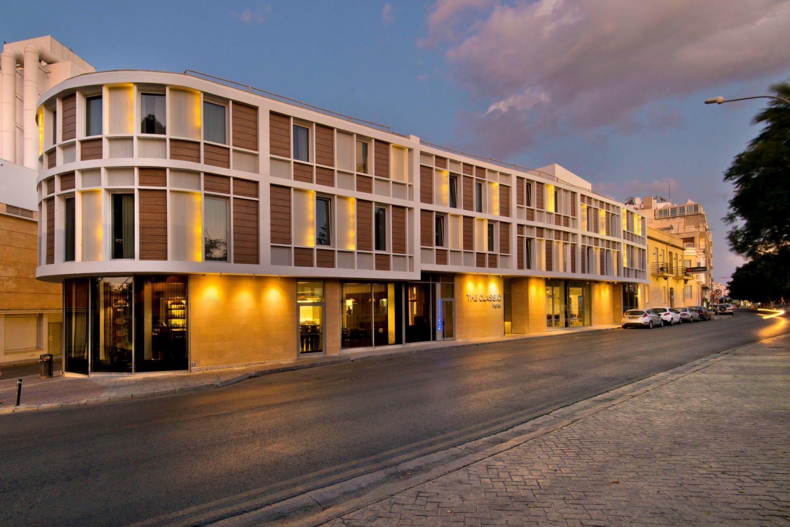 Modern hotel exterior on a city street at dusk with illuminated facade and entrance.