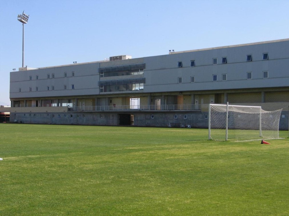 Grünes Fußballtrainingsfeld mit Torpfosten und modernem mehrstöckigem Sportgebäude