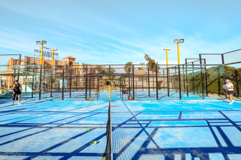 Frontalansicht benachbarter blauer Outdoor-Padelplätze mit spielenden Personen bei Tageslicht.