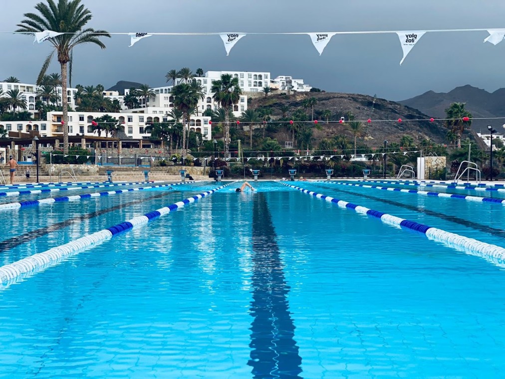Zentrierter Blick entlang eines Außen-Schwimmbeckens mit einem Schwimmer, Hanggebäuden und Palmen.