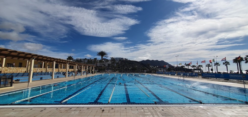 Weite Ansicht eines Außen-Schwimmbeckens mit Flaggen, Sitzbereichen und Hügeln unter teilweise bewölktem Himmel.