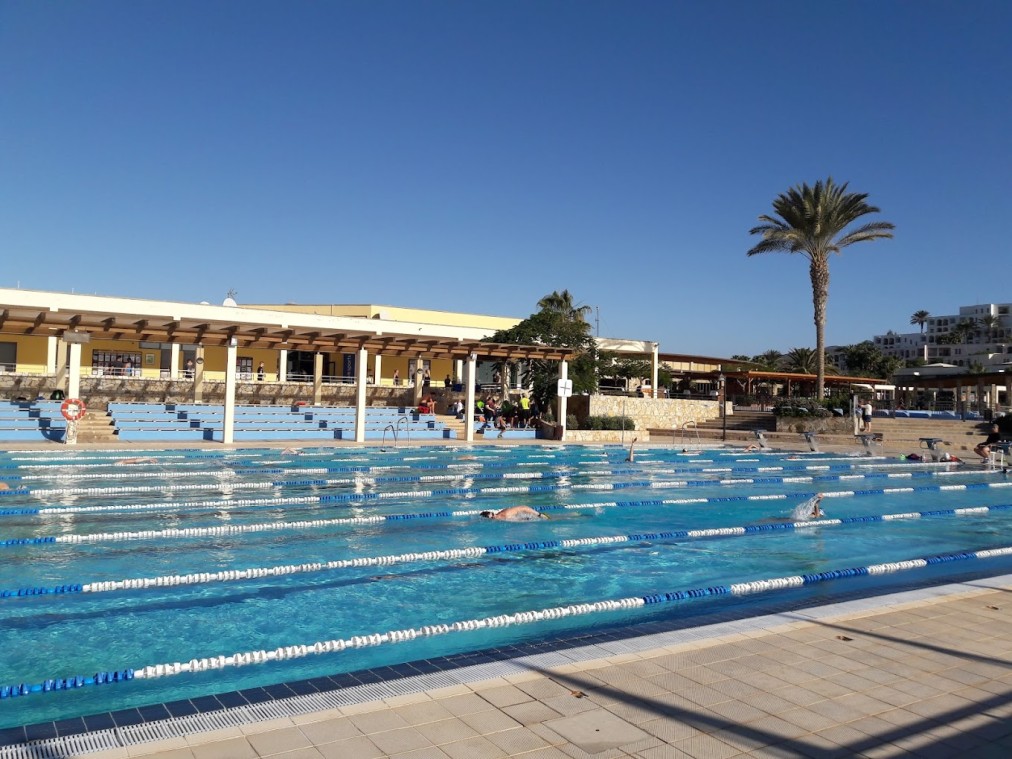 Athleten schwimmen Bahnen in einem Außen-Trainingsbecken neben schattigen Sitzplätzen und einem flachen Gebäude.