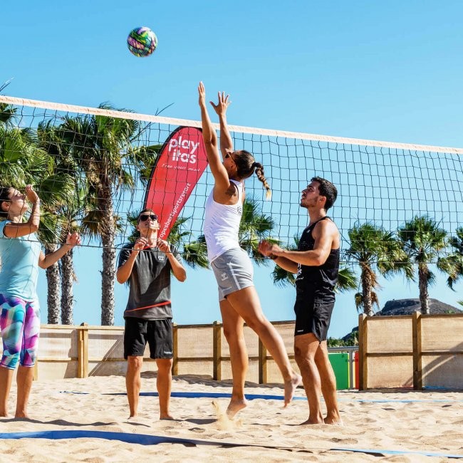 Beachvolleyballspiel auf einem Sandplatz mit Spielern am Netz und Palmen im Hintergrund.