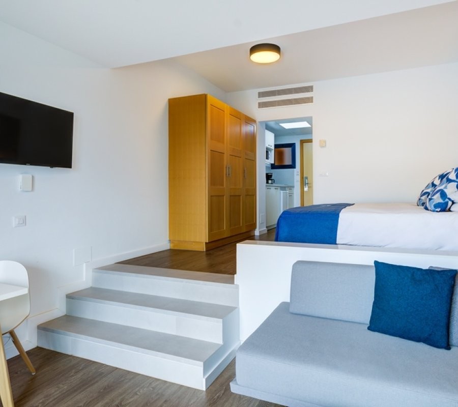 Studiozimmer mit erhöhtem Bettbereich, Sofa, Kleiderschrank und Zugang zur Küchenzeile.
