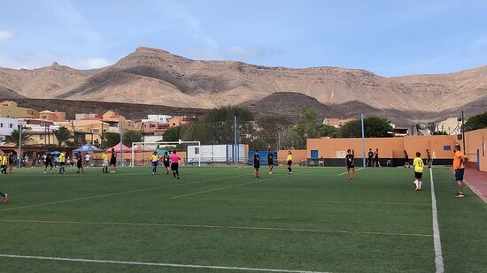 Fußballspiel oder Training auf einem eingezäunten Outdoor-Platz mit Bergkulisse und Teams auf dem Feld verteilt.