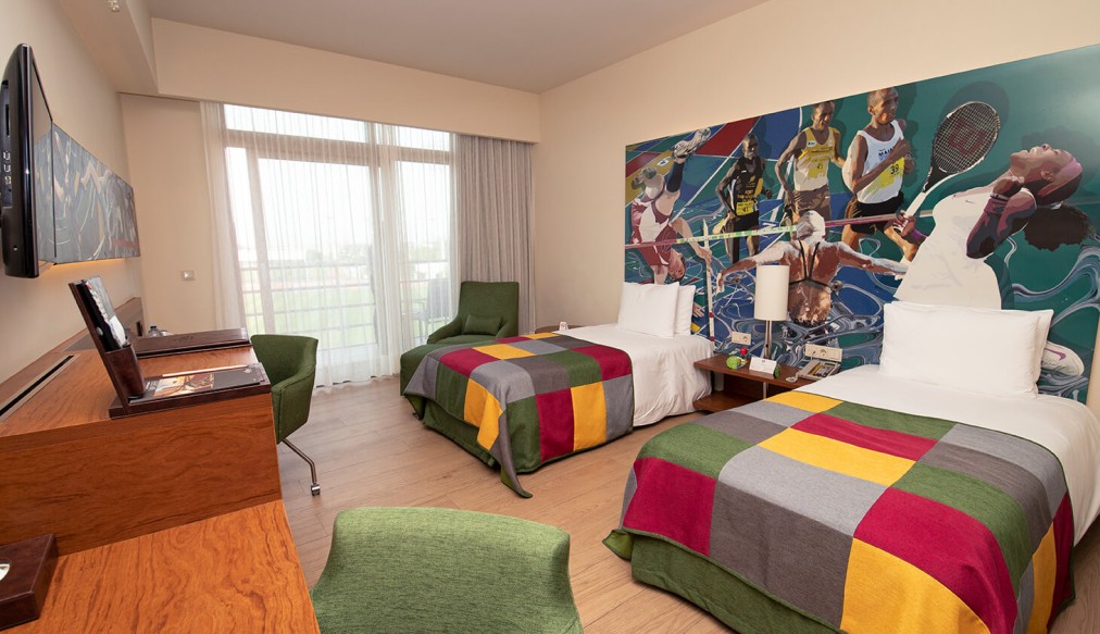 Großzügiges Zweibettzimmer mit zwei Betten, großem Sport-Mural und heller Fensterfront. Mit Schreibtisch, TV und Lounge-Sesseln – ideal für Sportler und Trainingsgruppen.