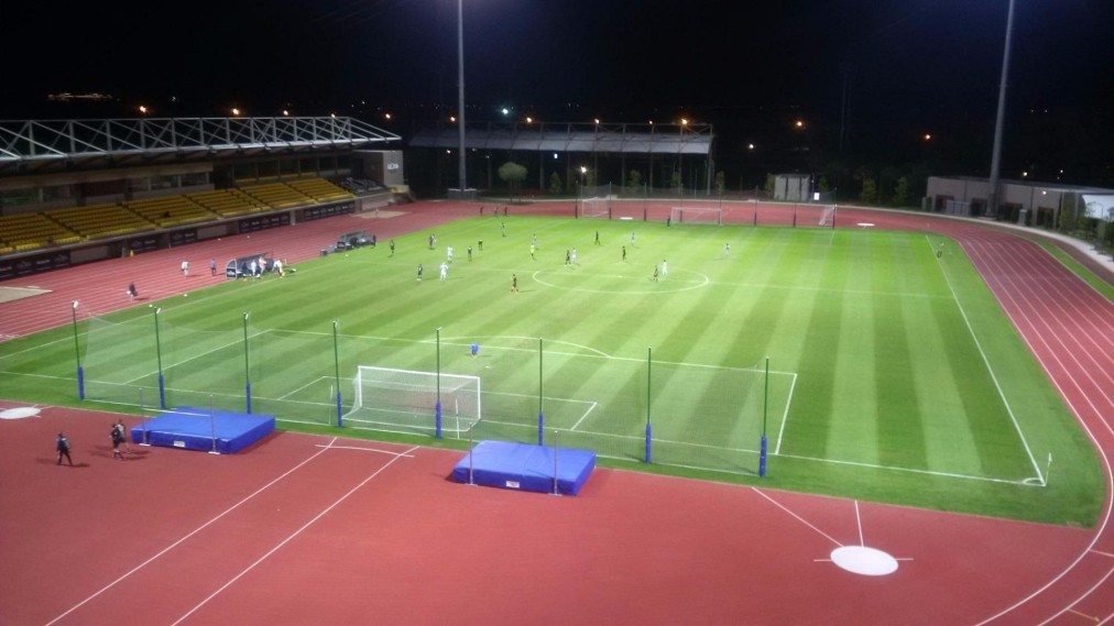 Flutlicht-Stadionansicht während eines Fußballspiels mit professioneller Wettkampf-Atmosphäre. Laufbahn und Tribünen bieten Raum für Events und Training bei Beleuchtung.