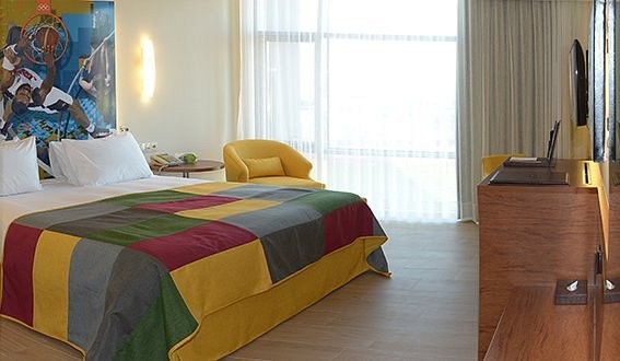 Helles Gästezimmer mit Doppelbett, farbenfroher Tagesdecke und sportlichem Wandmotiv. Ein großes Fenster sorgt für viel Tageslicht, ergänzt durch TV und einen Holzschreibtischbereich.