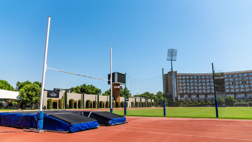 Leichtathletikbereich mit Hochsprungmatten neben Laufbahn und Rasenfeld auf einem Trainingscamp-Gelände.