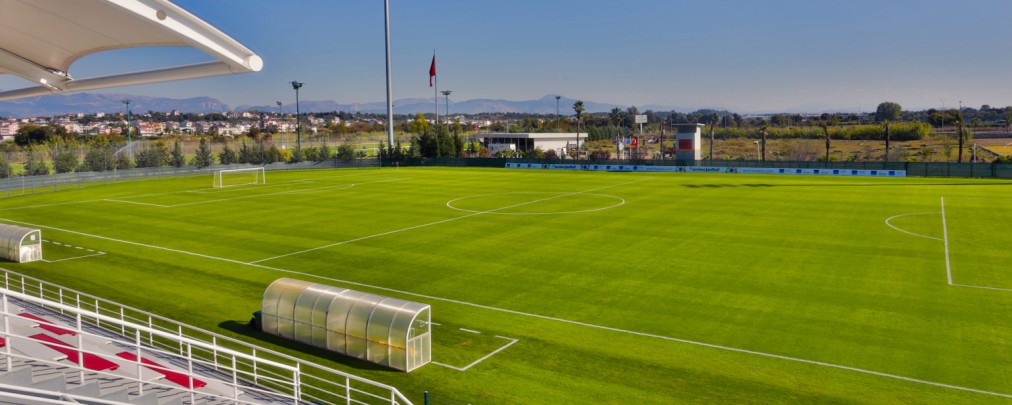 Fußballplatz in Originalgröße mit Teamkabinen und Flutlichtmasten, Blick von der Tribüne.