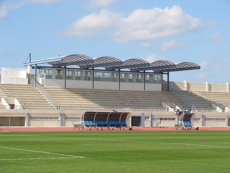 Fußballstadion mit Rasenplatz, Mannschaftsbänken und überdachter Haupttribüne.