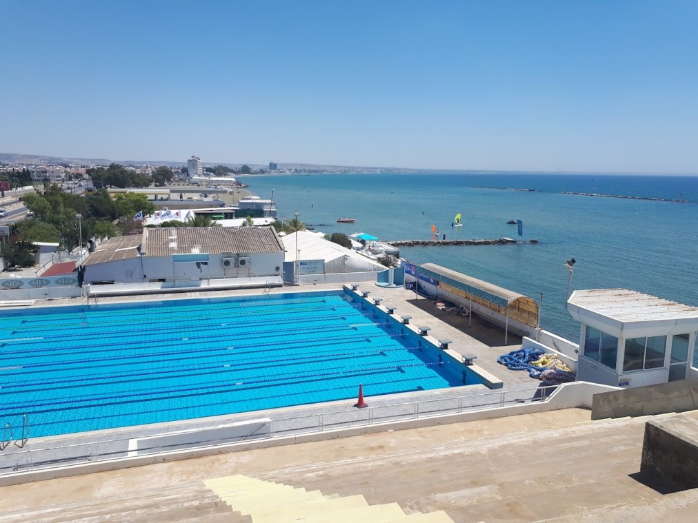 Außenliegendes Schwimmbecken am Meer mit Bahnenmarkierungen, Sitzplätzen am Beckenrand und Küstenblick im Hintergrund.