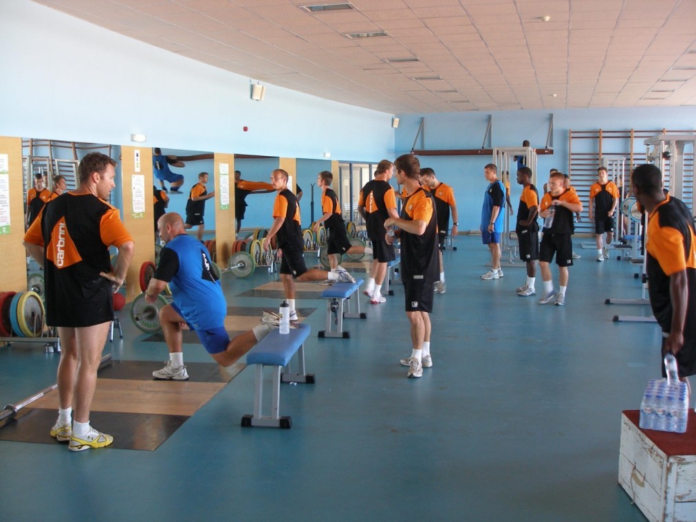 Athleten beim Training im Fitnessraum mit Bänken, Gewichten und Spiegeln.
