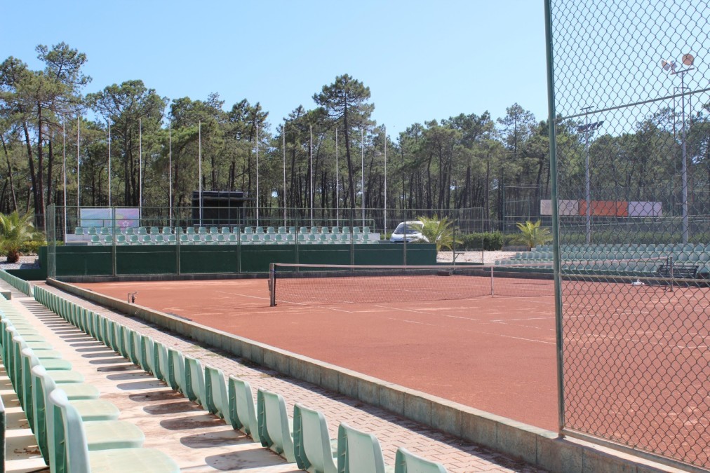 Tennis-Sandplatz mit Sitzreihen und Waldkulisse.
