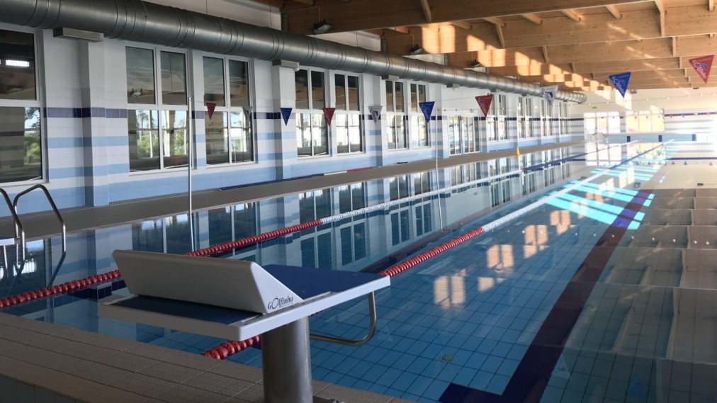 Leeres Hallenbad mit Schwimmleinen, einem Startblock und großen Fenstern.