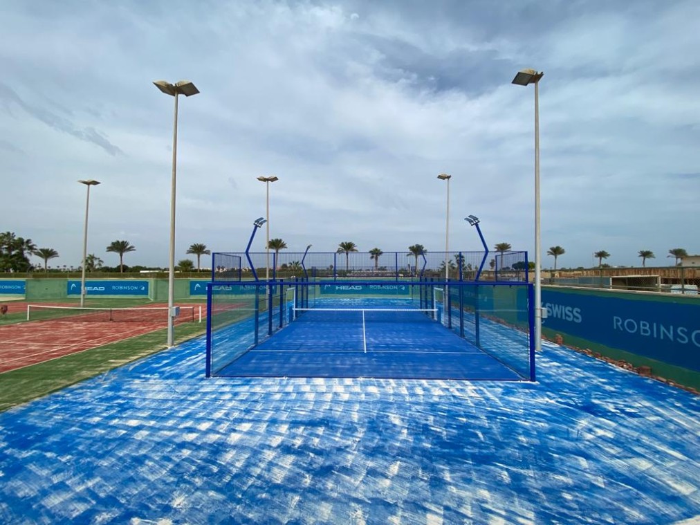 Leerer Outdoor-Padelcourt mit blauem Belag, Glasumrandung und hohen Flutlichtmasten.