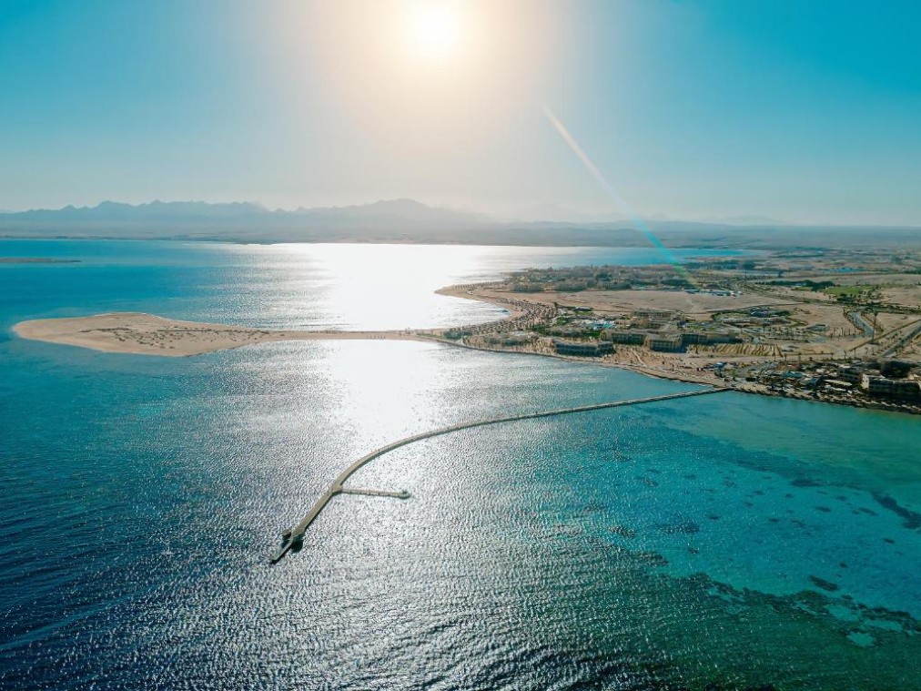 Luftaufnahme der Küste mit klarem blauen Meer, langem Pier, Sandstrand und Bergen im Hintergrund unter strahlender Sonne