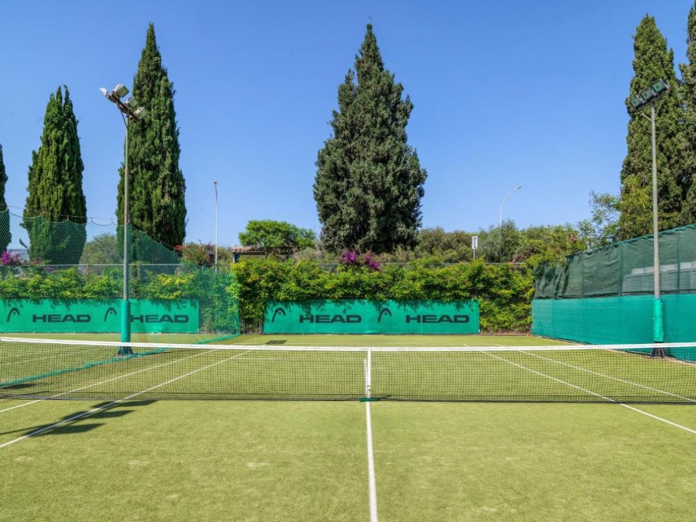 Außenliegender Tennisplatz mit grüner Oberfläche, umgeben von hohen Bäumen und grünen HEAD-Netzen für Trainingseinheiten