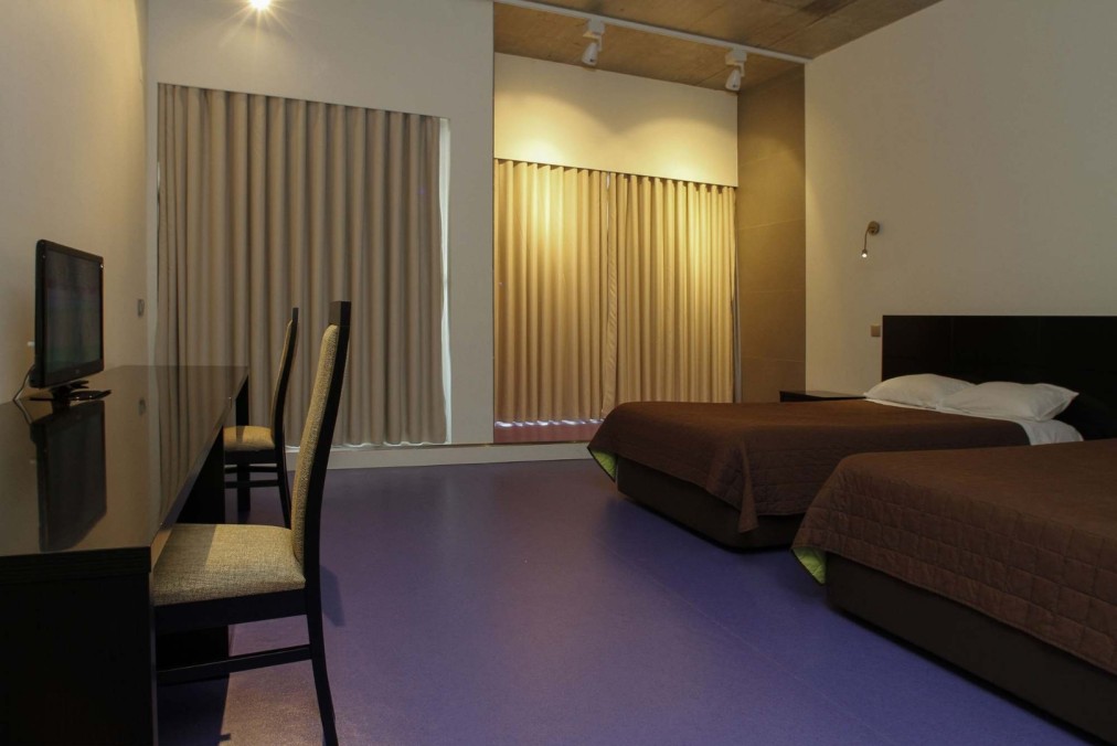 Schlichtes Zweibettzimmer mit zwei Einzelbetten, Schreibtisch und Fernseher sowie großen Fenstern mit Vorhängen.