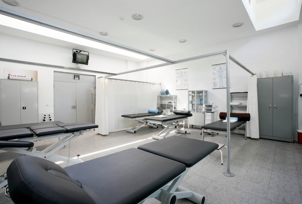 Heller Physiotherapie-Behandlungsraum mit mehreren Liegen, Sichtschutzvorhängen und medizinischen Schränken.