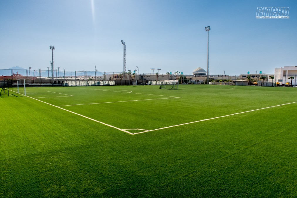 Weitwinkelansicht des grünen Fußballfelds im Trainingscamp mit hohen Flutlichtmasten und blauem Himmel