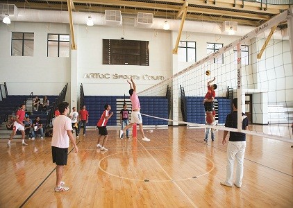 Hallen-Volleyballspiel auf Holzboden, Spieler springen am Netz, Zuschauer sitzen auf Bänken.