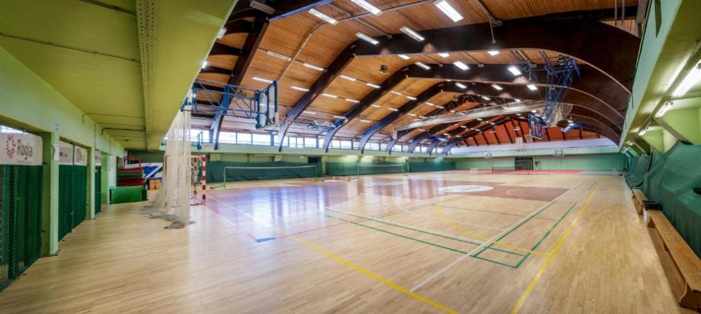 Indoor-Mehrzweck-Sporthalle mit Holzboden, Spielfeldmarkierungen und Basketballkörben.
