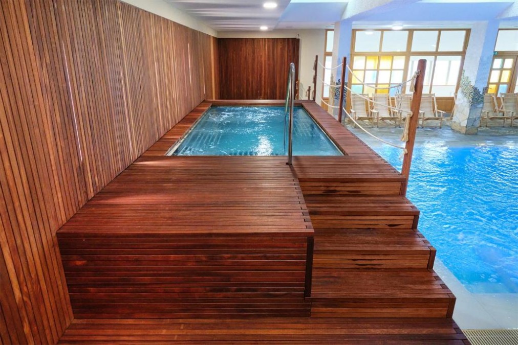 Hallenbad mit erhöhter Whirlpoolzone und Liegestühlen an großen Fenstern.