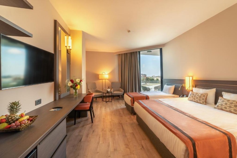 Helles Zweibettzimmer mit Schreibtisch, wandmontiertem TV, Sitzbereich und Balkontür.