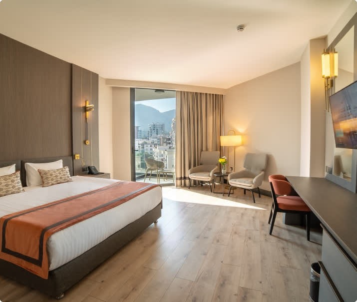 Modernes Hotelzimmer mit Doppelbett, Sitzecke, Schreibtisch und Balkonblick auf Stadt und Berge.