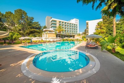 Außenpool mit Sonnenliegen und Sonnenschirmen, mit Blick auf Hotelgebäude und Gartenanlage.