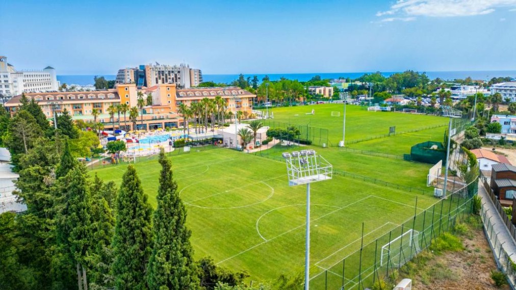 Luftaufnahme der Hotelanlage mit zwei Fußballplätzen und umliegenden Gebäuden nahe der Küste.