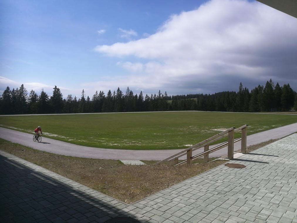 Rasenfußballplatz mit asphaltierter Runde, Waldhügeln und einem Radfahrer in der Nähe.