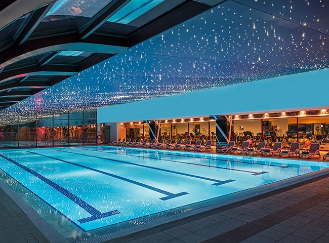 Weitere Ansicht des beleuchteten Innenpools mit Liegestühlen und dem Fitnessbereich hinter Glas.