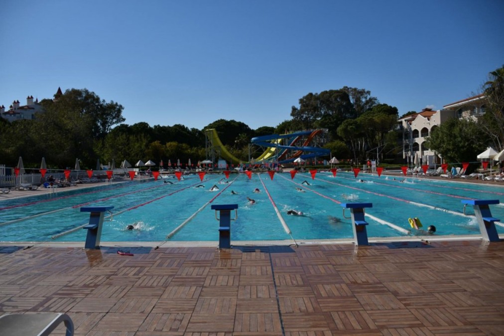 Außen-Schwimmbecken mit Bahnen, Startblöcken und Schwimmern; dahinter Hotelgebäude und Wasserrutschen.