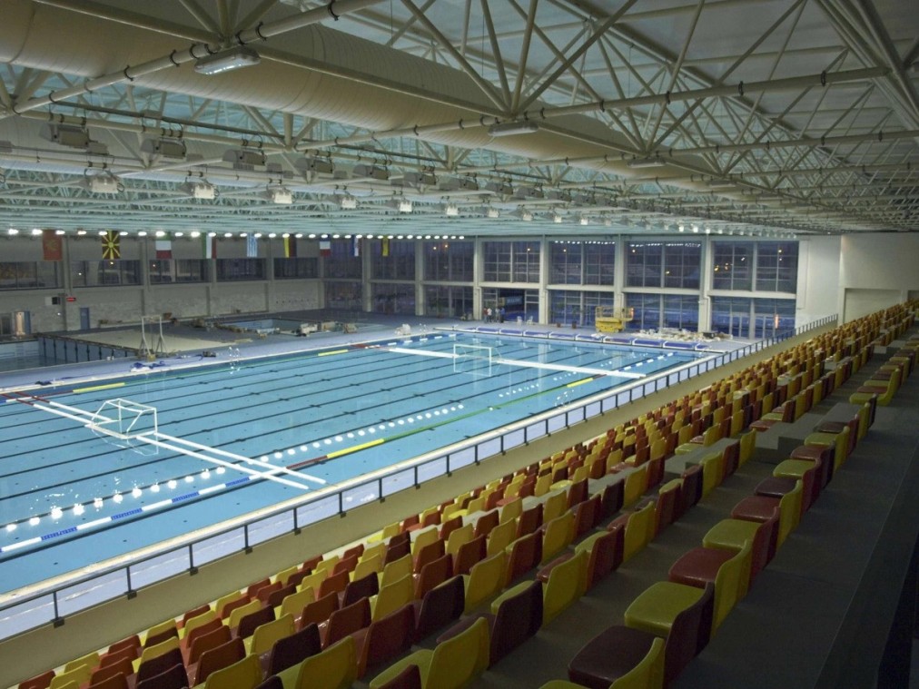 Weite Ansicht eines Hallenbeckens für Wasserball mit Toren und Zuschauerplätzen.