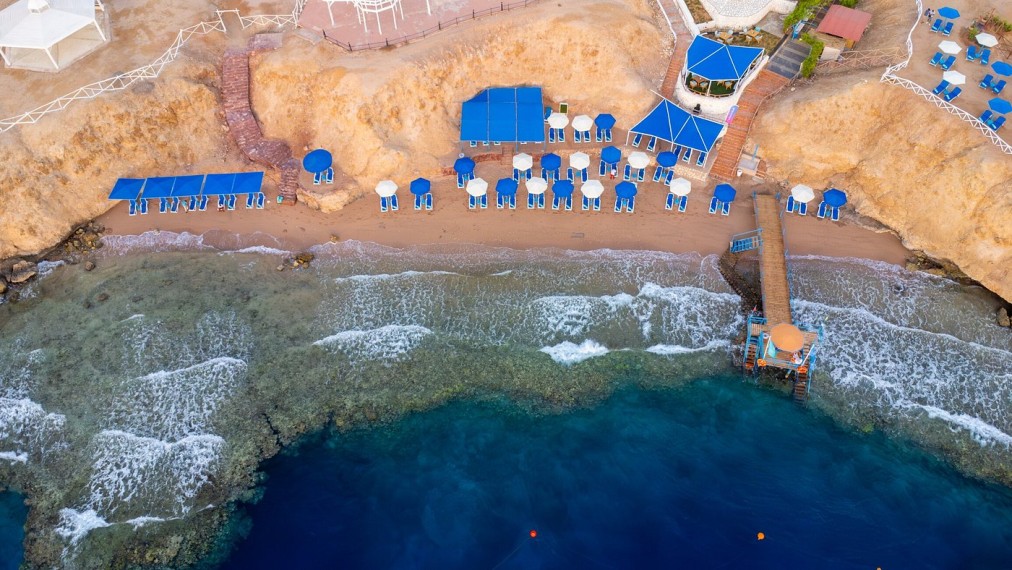 Luftaufnahme eines Privatstrandes mit blauen Sonnenschirmen, Liegestühlen und einem Holzsteg über klarem Wasser