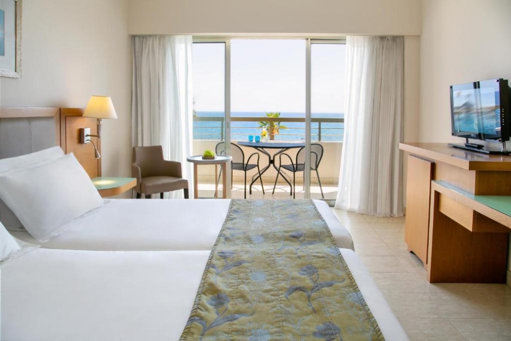 Gästezimmer mit Meerblick-Balkon, zwei Betten, TV und Schreibtischbereich.