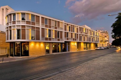 Moderne Hotelaußenansicht an einer Stadtstraße bei Dämmerung mit beleuchteter Fassade und Eingang.