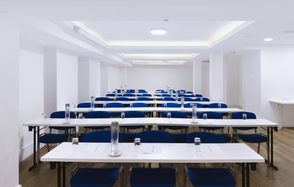 Konferenzraum mit Tischen im Schulungsstil, blauen Stühlen, Wasserflaschen und Notizblöcken.