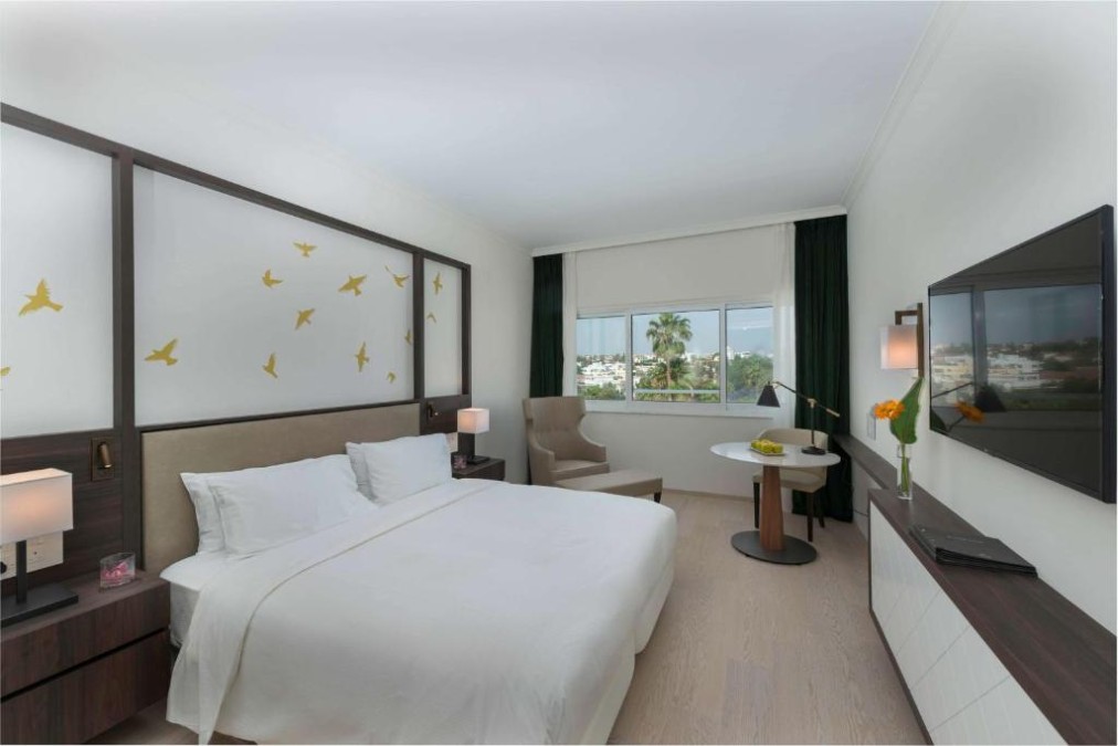 Zimmer mit Kingsize-Bett, Wandfernseher, Tisch und Stadtblick.