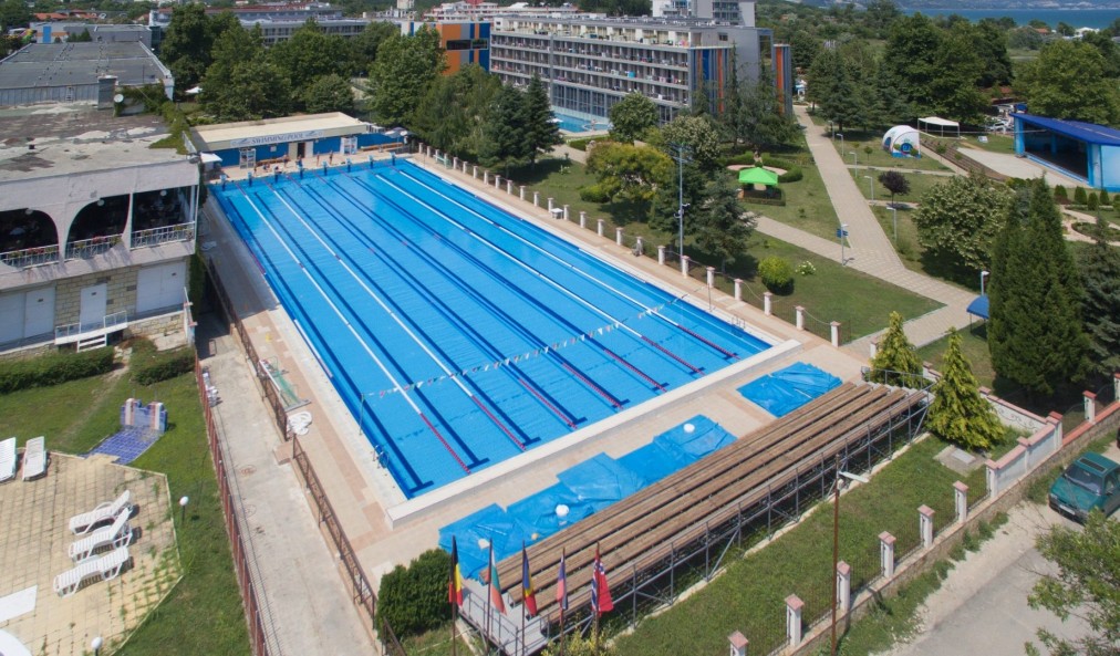 Grande piscine olympique extérieure bleue avec marquages de couloirs et fanions, située parmi les arbres pour le stage d'entraînement des athlètes