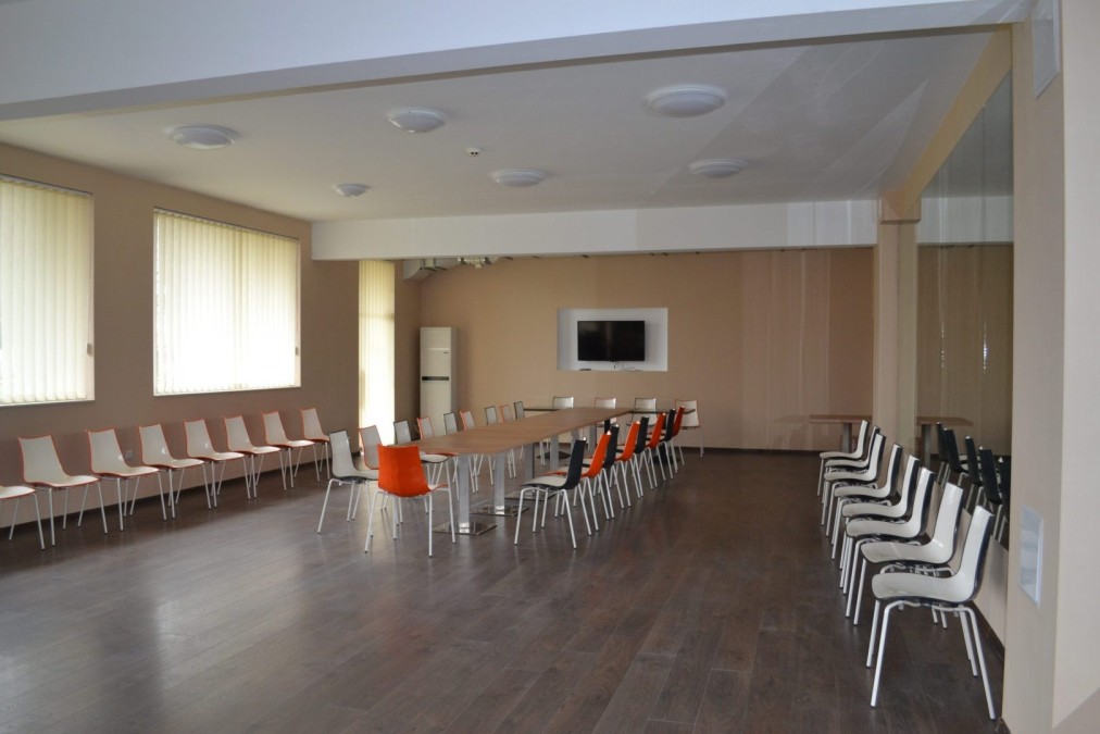 Grande salle polyvalente avec parquet, chaises et écran, prête pour les réunions de coaching ou briefings
